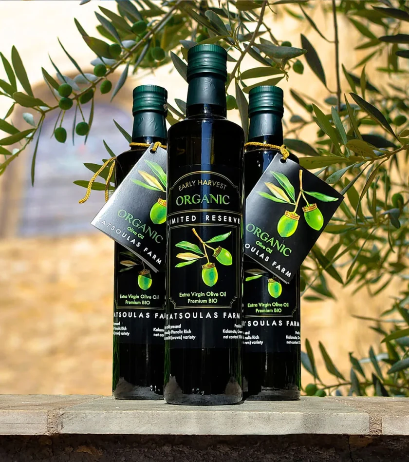 Three bottles of Katsoulas Farm olive oil with black labels and tags, among olive branches