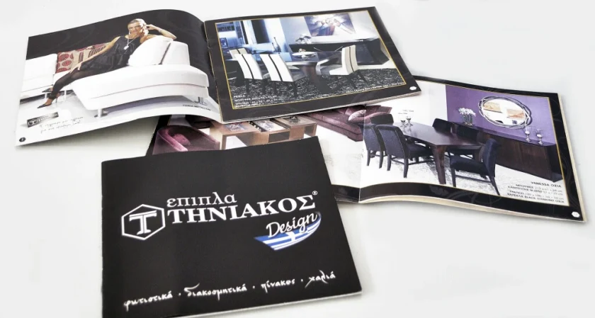 Το μαύρο εξώφυλλο του καταλόγου Έπιπλα Τηνιακός Design δίπλα σε ανοιχτές σελίδες