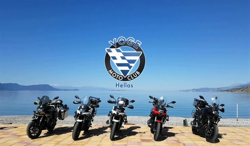 Πέντε μοτοσυκλέτες σε παραλία, με το λογότυπο του Voge Moto Club Hellas πάνω από τη θάλασσα